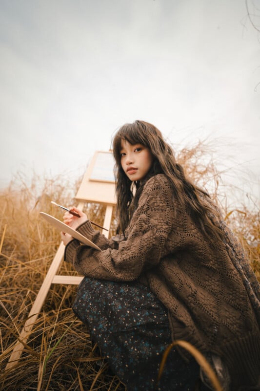 A young woman with long dark hair, wearing a brown sweater and patterned skirt, sits in a field of tall dry grass, sketching on a pad in front of a wooden easel under a cloudy sky.