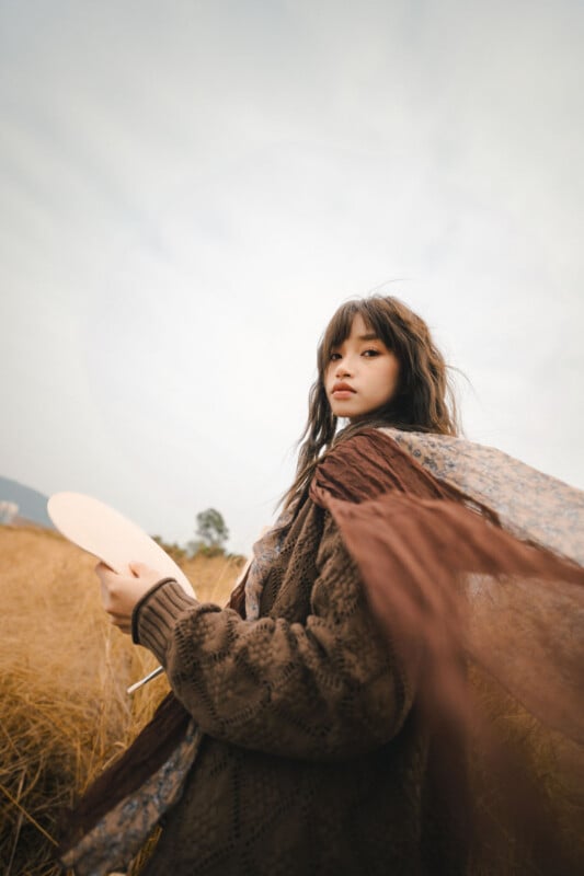 A young woman with long hair stands in a field of tall grass, wearing a brown knit sweater and a flowing scarf. She holds a round palette in one hand and gazes thoughtfully at the camera under a cloudy sky.