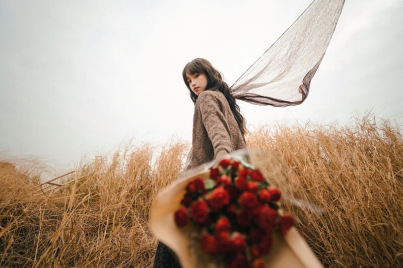 A woman in a brown outfit stands in a dry grassy field, holding a bouquet of red flowers toward the camera. A sheer brown scarf trails behind her in the wind against a cloudy sky.