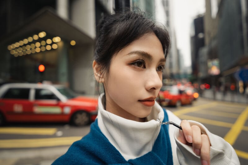 A woman with long dark hair and natural makeup stands on a city street, wearing a blue and white zip-up top. Behind her, red taxis and buildings line the busy urban scene.