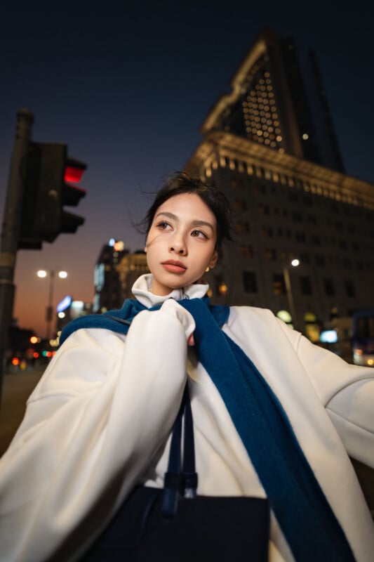 A young woman in a white coat and blue scarf stands on a city street at dusk, with tall buildings and city lights in the background. She looks thoughtful, gazing off to the side.