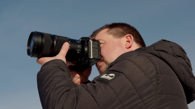 A person in a black jacket holds a camera up to their face, looking through the viewfinder and taking a photo against a clear blue sky.
