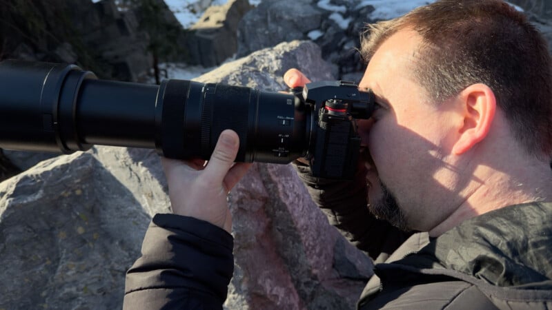 A person with short hair and a beard is outdoors, holding a camera with a large telephoto lens, aiming it towards the left. Rocks and some snow are visible in the background.