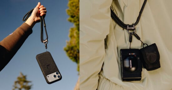 A hand holds a smartphone on a black lanyard against the sky, while another lanyard secures a camera and small pouch to a person in a light-colored jacket.