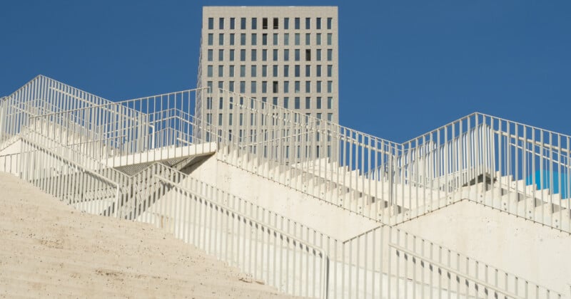 A modern, angular white staircase with metal railings rises sharply in the foreground, while a rectangular gray building with a grid of windows stands against a clear blue sky in the background.