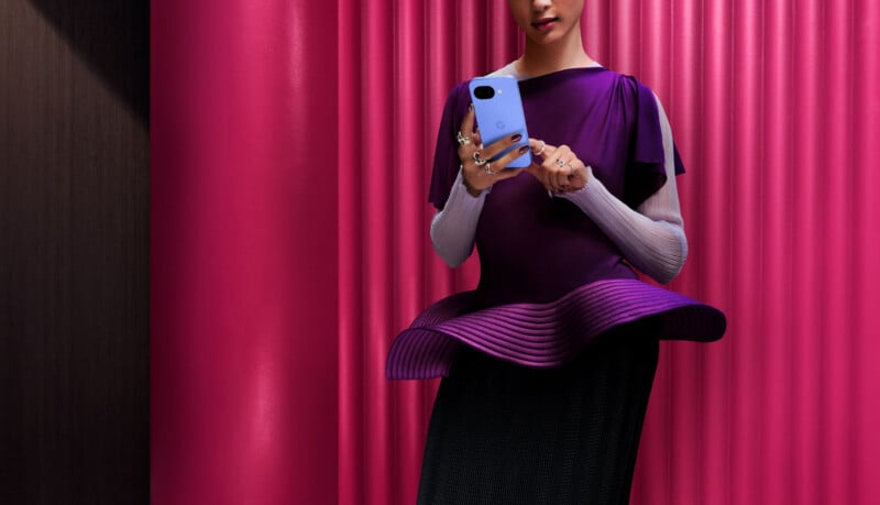 A person wearing a stylish, futuristic purple outfit stands in front of bright pink vertical panels, holding and looking at a blue smartphone. Their face is partially visible.