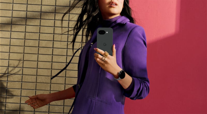 A woman in a purple outfit stands against a wall with tan brick and a pink section, holding a black smartphone and wearing several rings and a smartwatch. Her face is partially visible.