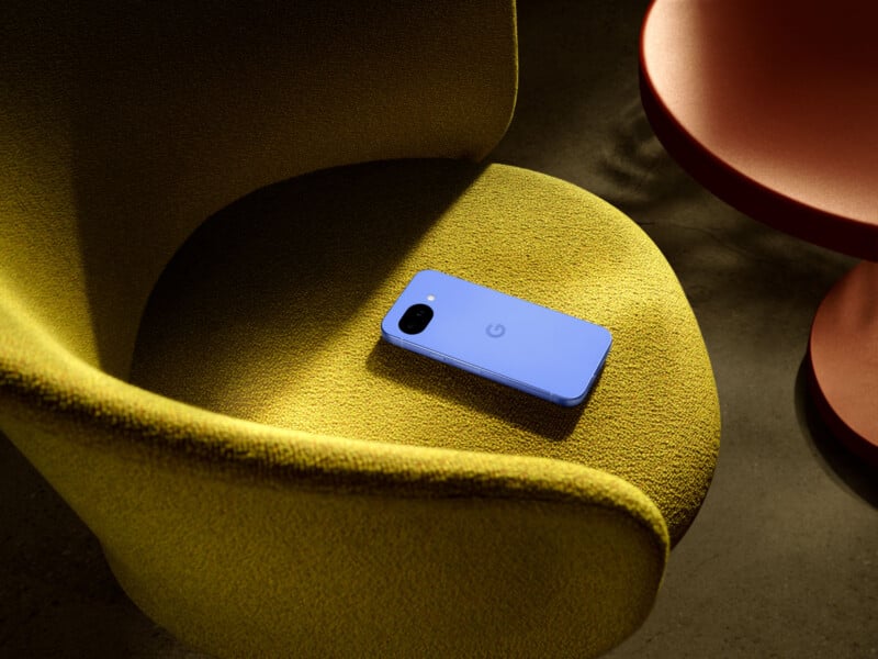 A blue smartphone rests face down on a textured, yellow-green upholstered chair next to a round, orange table.