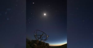 A metal sculpture shaped like a sphere stands on stone blocks under a night sky filled with stars, with a bright moon glowing near the center and a faint glow on the horizon.