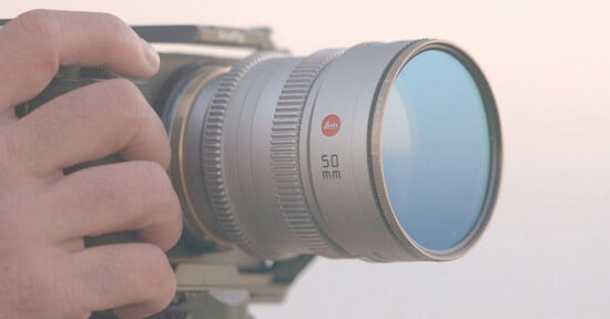 A close-up of a hand adjusting a camera with a silver Leica 50mm lens, set against a soft, light background.