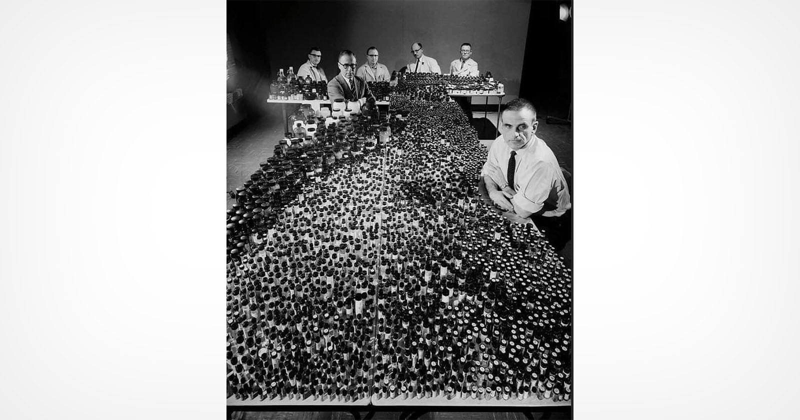 Five scientists sit behind long tables crowded with hundreds of small glass bottles, possibly used for research or experiments, in a black-and-white photo. One man stands at the front, looking toward the camera.