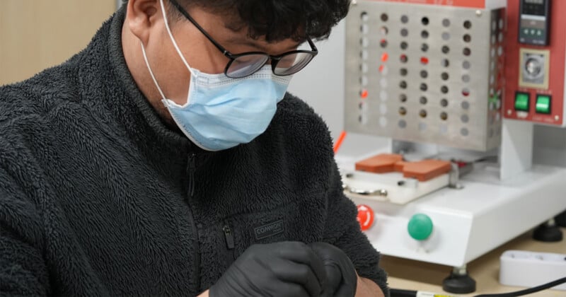 A person wearing glasses, a blue surgical mask, and black gloves works with their hands near laboratory equipment, including a machine with buttons and dials, in a lab or workshop setting.