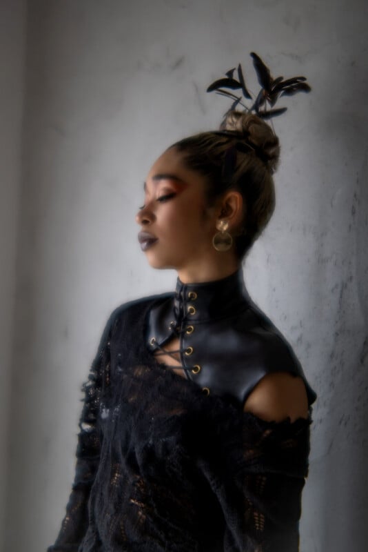 A person with dramatic makeup, dark lipstick, and wearing a black lace outfit with leather accents poses against a textured gray wall. Their hair is styled in a high bun adorned with black decorative elements.