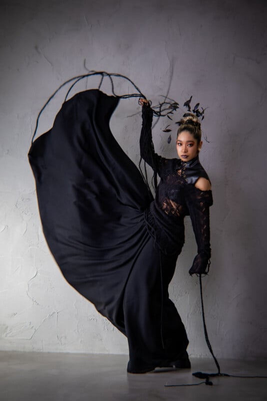 A woman in an elaborate black dress poses gracefully with one leg raised, creating dramatic movement in the skirt. She wears a black lace top and a decorative headpiece, standing against a light textured wall.