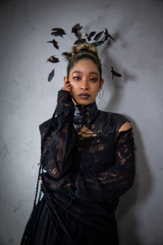 A woman in a black, lace Gothic outfit poses against a gray textured wall. She has dark makeup, wears leaf-like headpieces, and gazes confidently at the camera with one hand touching her face.