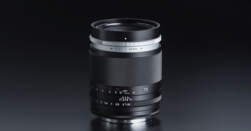A close-up of a black camera lens with white and red markings, silver accents, and engraved numbers, placed upright on a reflective dark surface against a black background.