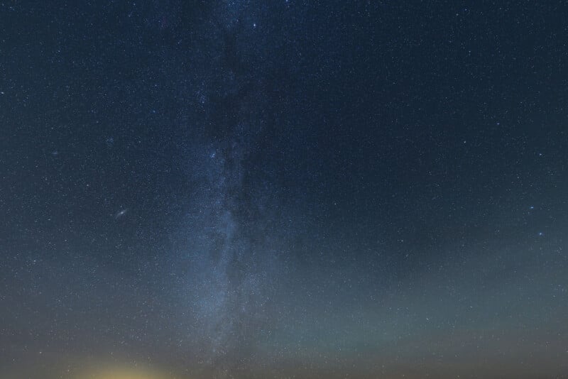A clear night sky filled with countless stars and a dense, faint band of the Milky Way stretching vertically across the image above a dark horizon.