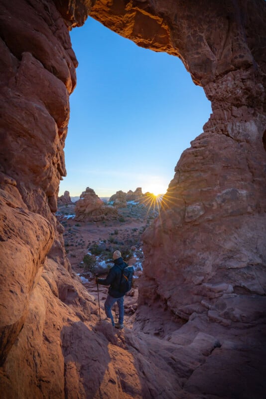 A hiker with a backpack and walking stick stands between large red rock formations, looking out at a desert landscape as the sun rises on the horizon, casting a warm glow over the scene.
