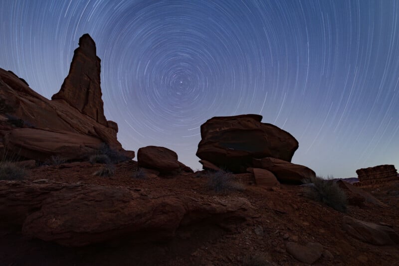 Rocky desert landscape at night with a tall eroded rock formation; long-exposure photo shows circular star trails in the clear sky above the scene.