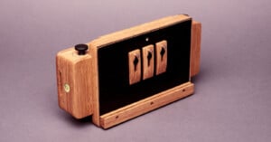 A handcrafted wooden camera with black panels and three toggle switches on the front, placed on a plain background. The design combines natural wood with functional hardware elements.