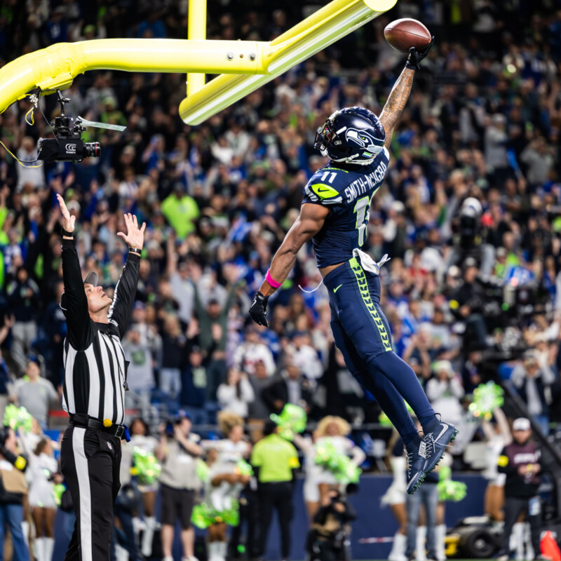A Seattle Seahawks player leaps and dunks a football over the goalpost while a referee raises both arms, with a cheering crowd in the background.