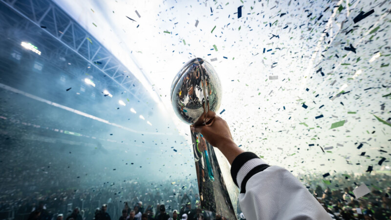 A person holds up the Vince Lombardi Trophy as confetti falls in a packed stadium, celebrating a Super Bowl victory.