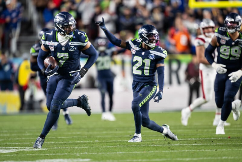 Seattle Seahawks football players in navy uniforms run down the field during a game. One player holds the ball while teammates run alongside, and opposing team members in white are seen in the background.