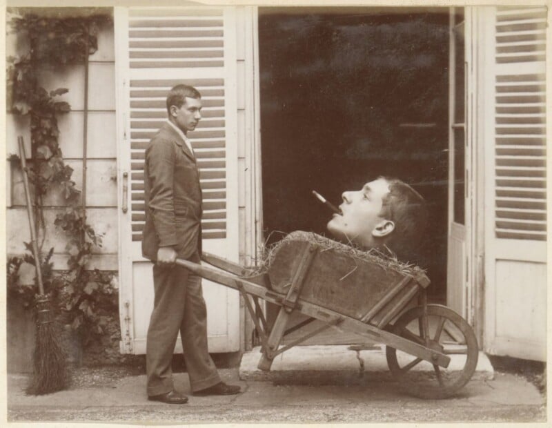 A man in a suit stands outside with a wooden wheelbarrow filled with hay. Among the hay is a large, surreal human head with a cigarette in its mouth. Shutters and a broom are visible in the background.