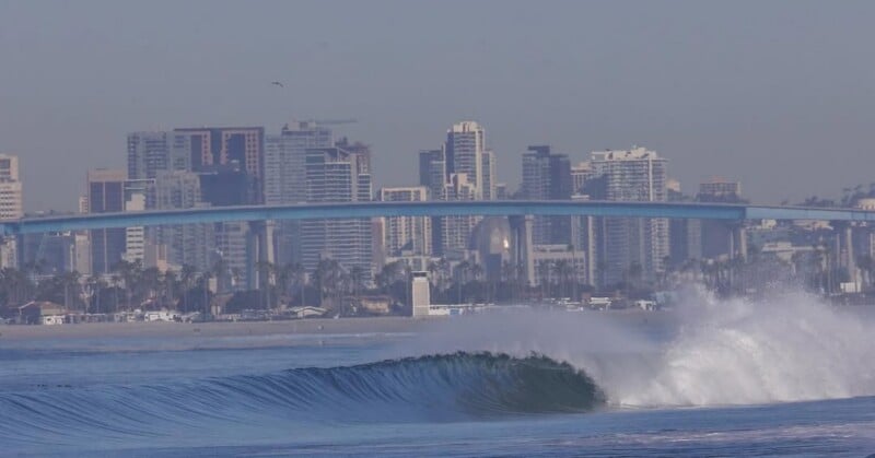 A breaking ocean wave in the foreground with a city skyline and a long, blue bridge in the background under a clear sky.