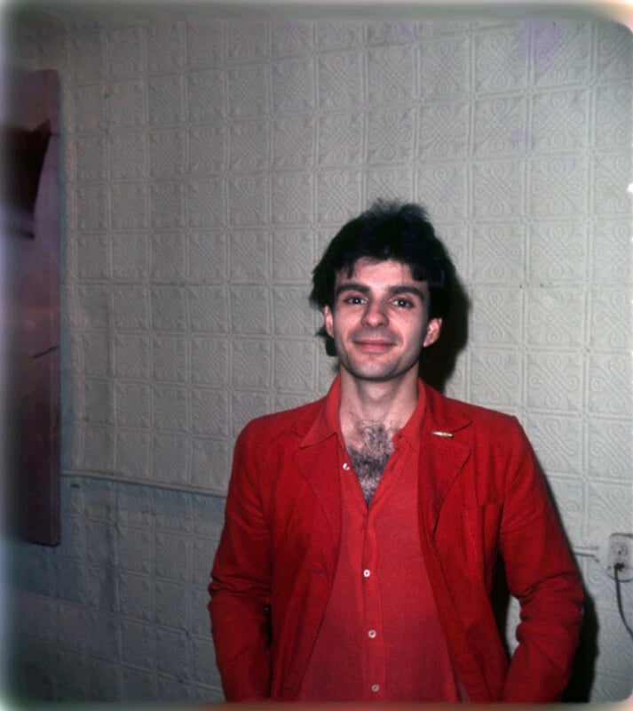A person with dark hair, wearing a red blazer over a red shirt, stands and smiles in front of a patterned beige wall.