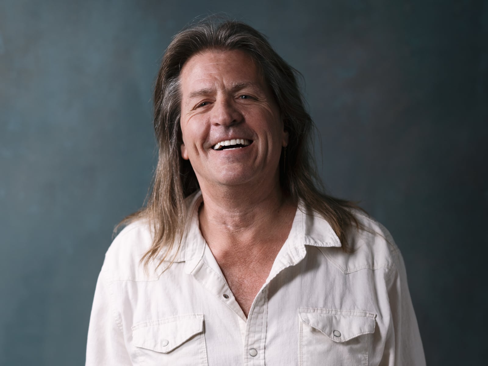 A middle-aged man with long brown hair smiles warmly, wearing a cream-colored button-up shirt against a dark, softly blurred background.