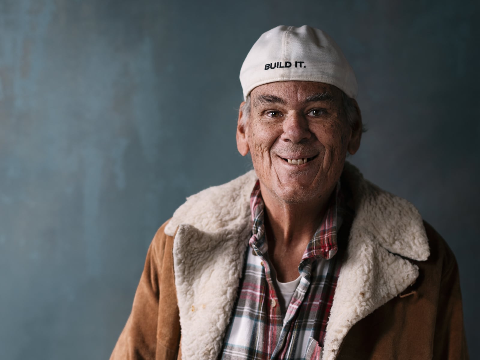 An older man wearing a white cap backward, brown jacket with a shearling collar, and plaid shirt smiles warmly in front of a blue textured background.
