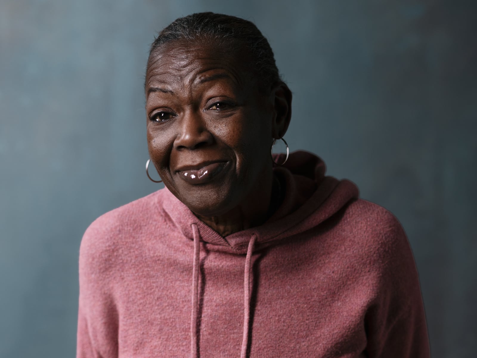 An older woman with short hair and hoop earrings wears a pink hoodie and smiles gently at the camera, standing against a softly blurred blue-gray background.
