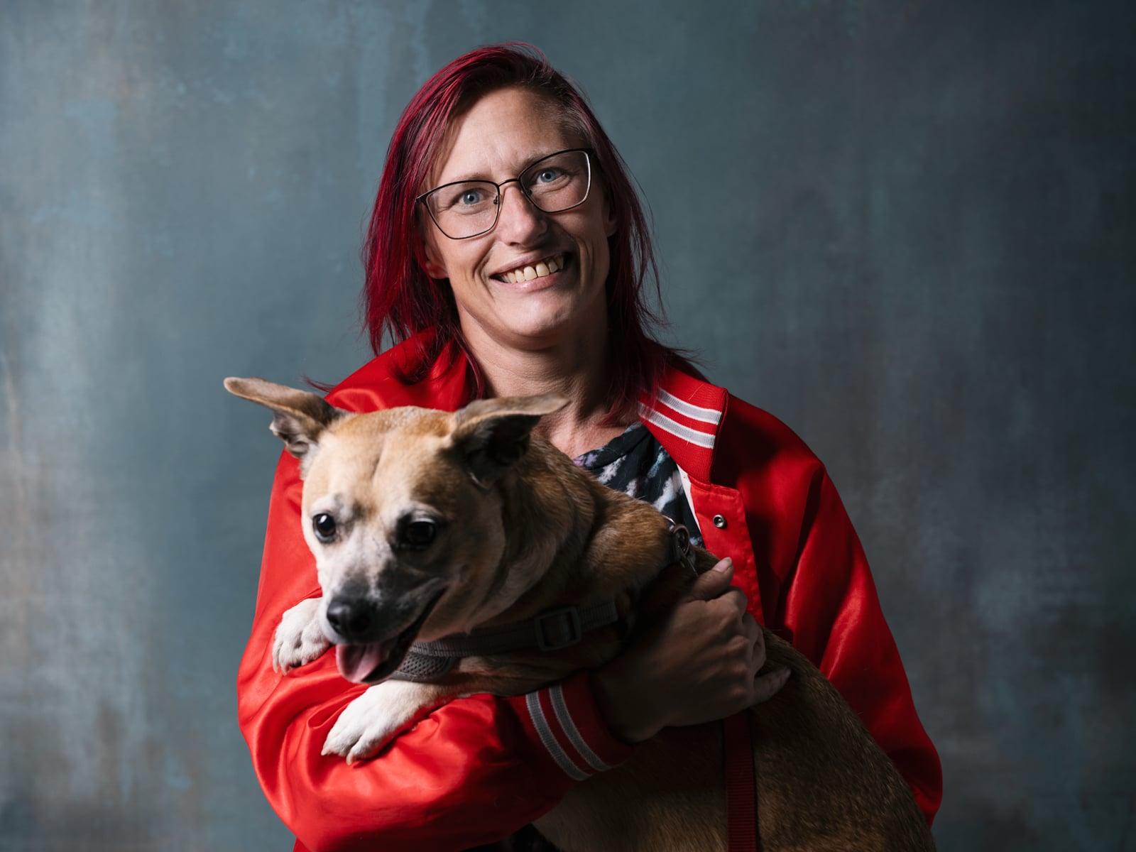 A person with red hair and glasses, wearing a red jacket, smiles while holding a small tan and white dog against a blue-gray background.
