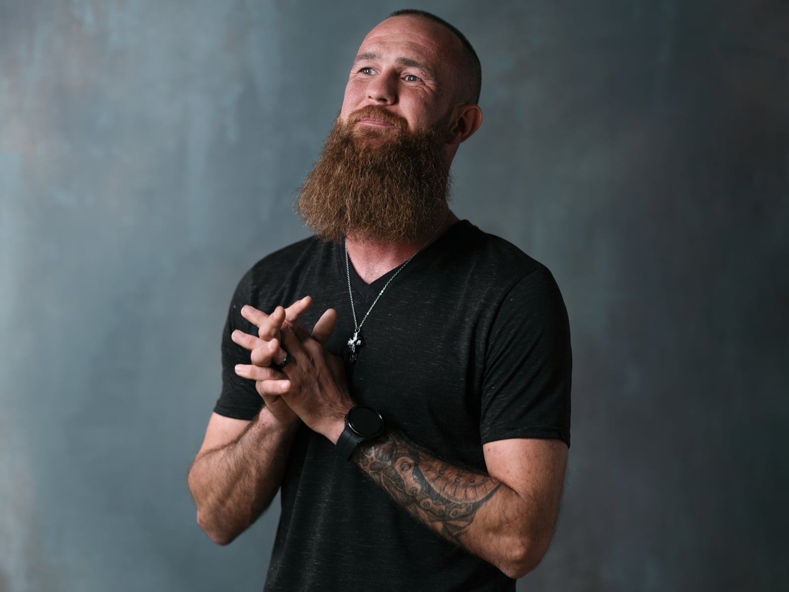 A bearded man with a tattooed arm stands against a gray background, wearing a black t-shirt, a necklace with a cross, and a watch. He clasps his hands and looks upward with a thoughtful expression.