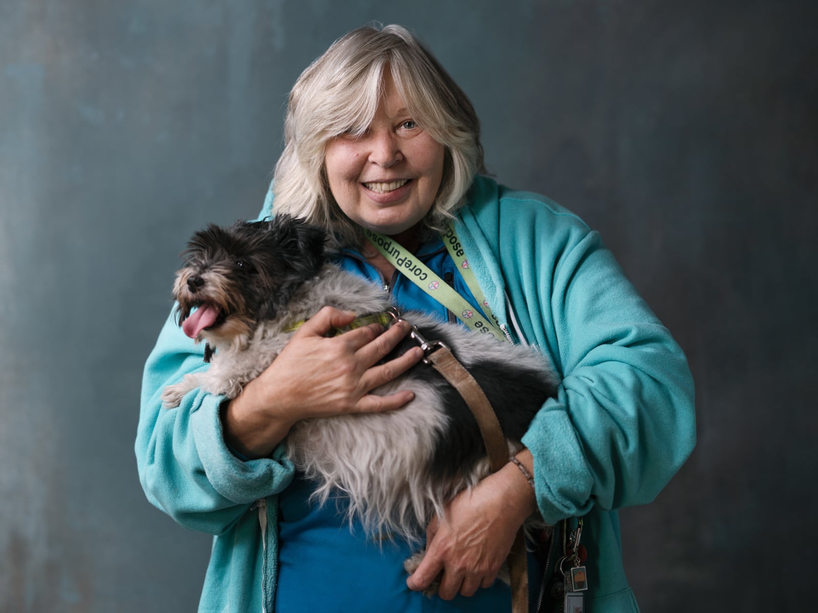 A smiling woman with gray hair wearing a teal jacket holds a small, fluffy black and white dog. The dog is panting happily, and the woman wears a colorful lanyard around her neck against a dark background.