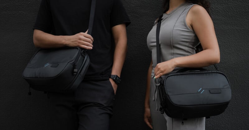 Two people standing side by side against a dark wall, each wearing a crossbody black bag. The person on the left wears a black outfit, while the person on the right wears a gray jumpsuit. Both hold their bags with one hand.