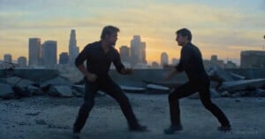 Two men wearing dark clothing face each other in a fighting stance on a rubble-strewn rooftop with a city skyline and sunset in the background. The image is labeled as AI generated.