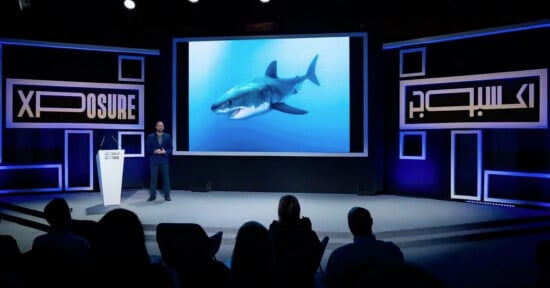 A speaker stands on stage beside a large screen displaying a great white shark underwater, with an audience seated in front. The stage is lit in blue with "Xposure" signage on both sides.