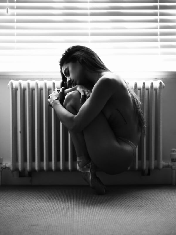 En una fotografía en blanco y negro, una mujer sentada desnuda en posición fetal en el suelo frente a un radiador, iluminada por persianas.