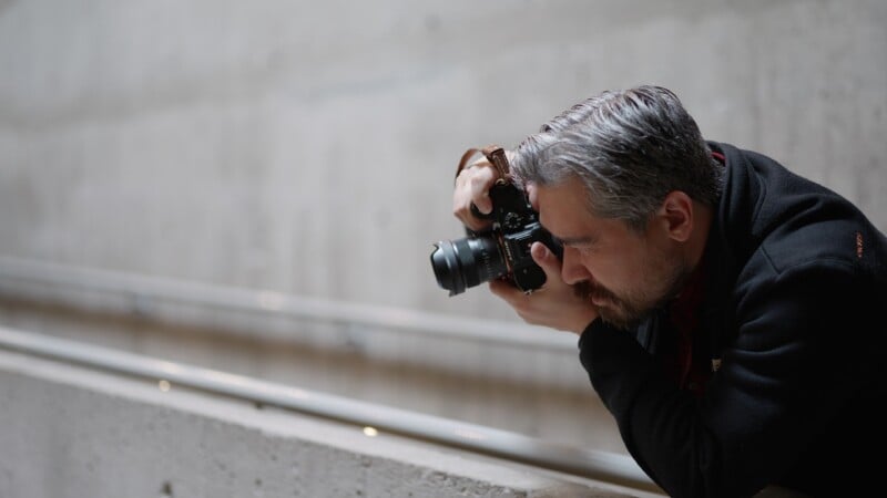 A man with gray hair and a beard leans over a concrete railing, intently looking through a camera as he takes a photo. The background is blurred and consists of plain, gray concrete walls.