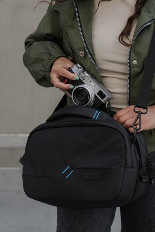 A person in a green jacket holds a silver camera and a black shoulder bag with blue accents, preparing to place the camera inside the bag.