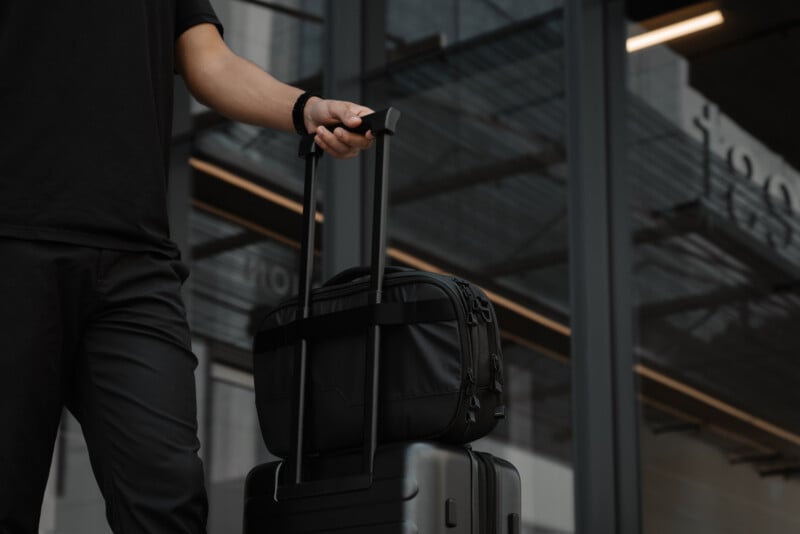 A person dressed in black pulls a black suitcase with an attached bag outside a modern building with large glass windows.
