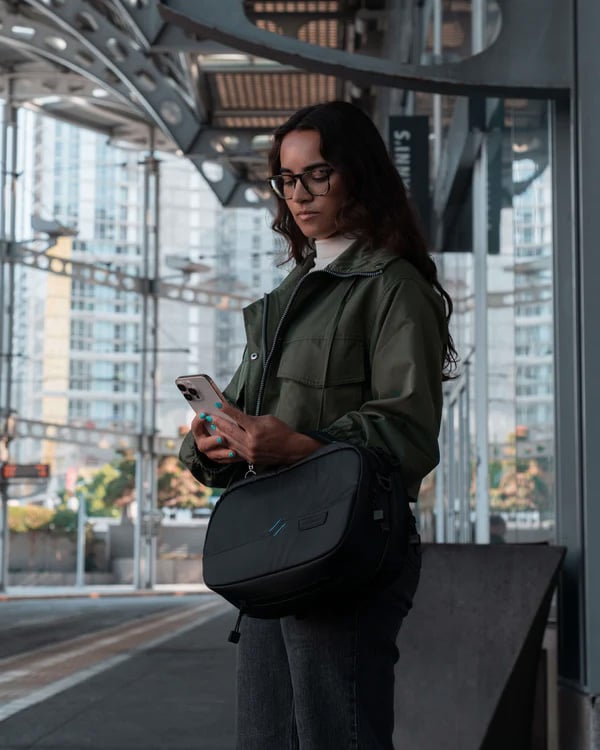 A woman wearing glasses and a green jacket stands at an outdoor transit station, looking at her phone and holding a black bag across her body. City buildings and metal structures are visible in the background.