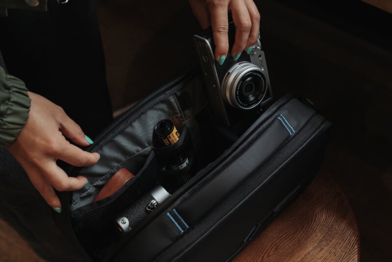 A person with teal-painted nails places a camera into a black camera bag, which also contains a lens and other accessories, on a wooden surface.