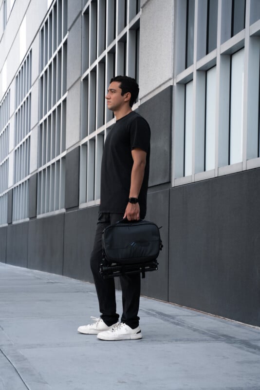 A person in black clothing and white sneakers stands on a sidewalk beside a modern building with large windows, holding a black bag in one hand and facing to the left.