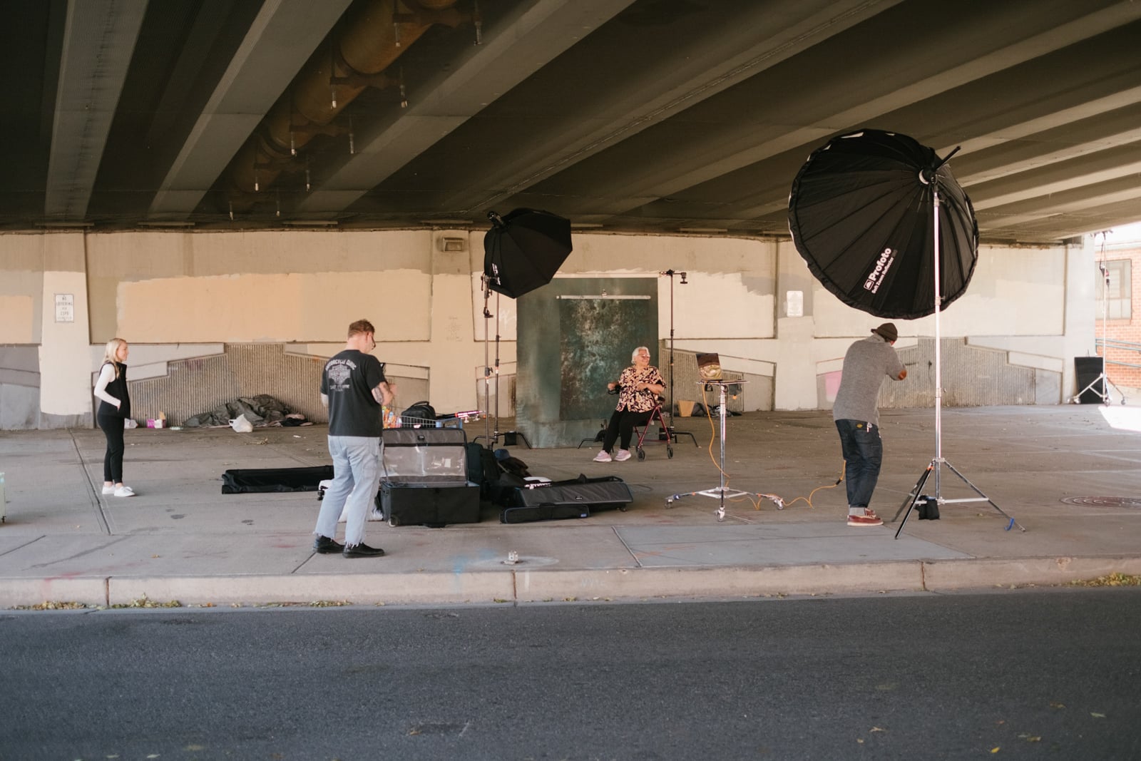 A photography crew sets up lighting equipment under a bridge, preparing to shoot a woman seated in the center. Three crew members are adjusting equipment, while bags and supplies are scattered on the ground nearby.