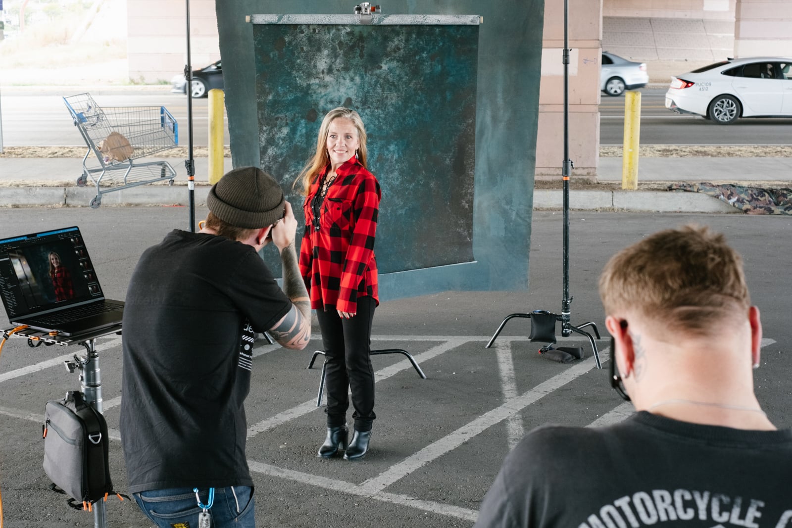 A woman in a red plaid shirt poses for a portrait in front of a backdrop outdoors, while a photographer takes her photo and another person stands nearby. A shopping cart and cars are visible in the background.