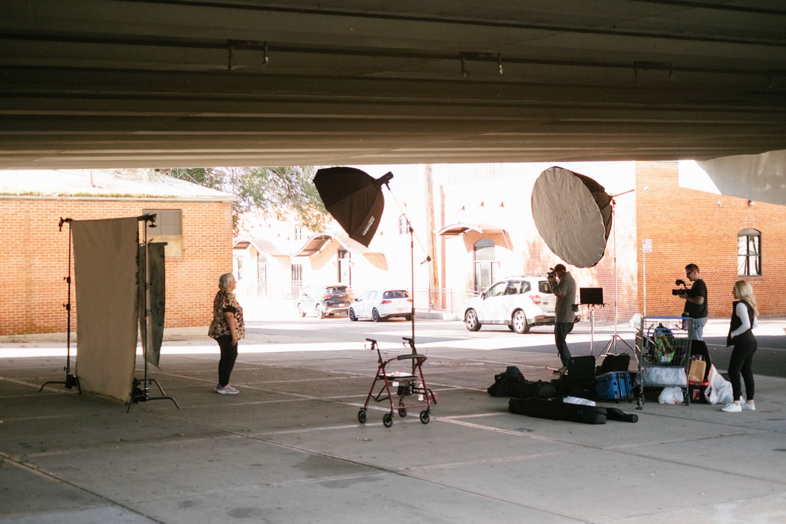 A woman stands in front of a backdrop under an overpass while a photographer and two assistants set up lighting and equipment; a walker and bags are on the ground nearby.