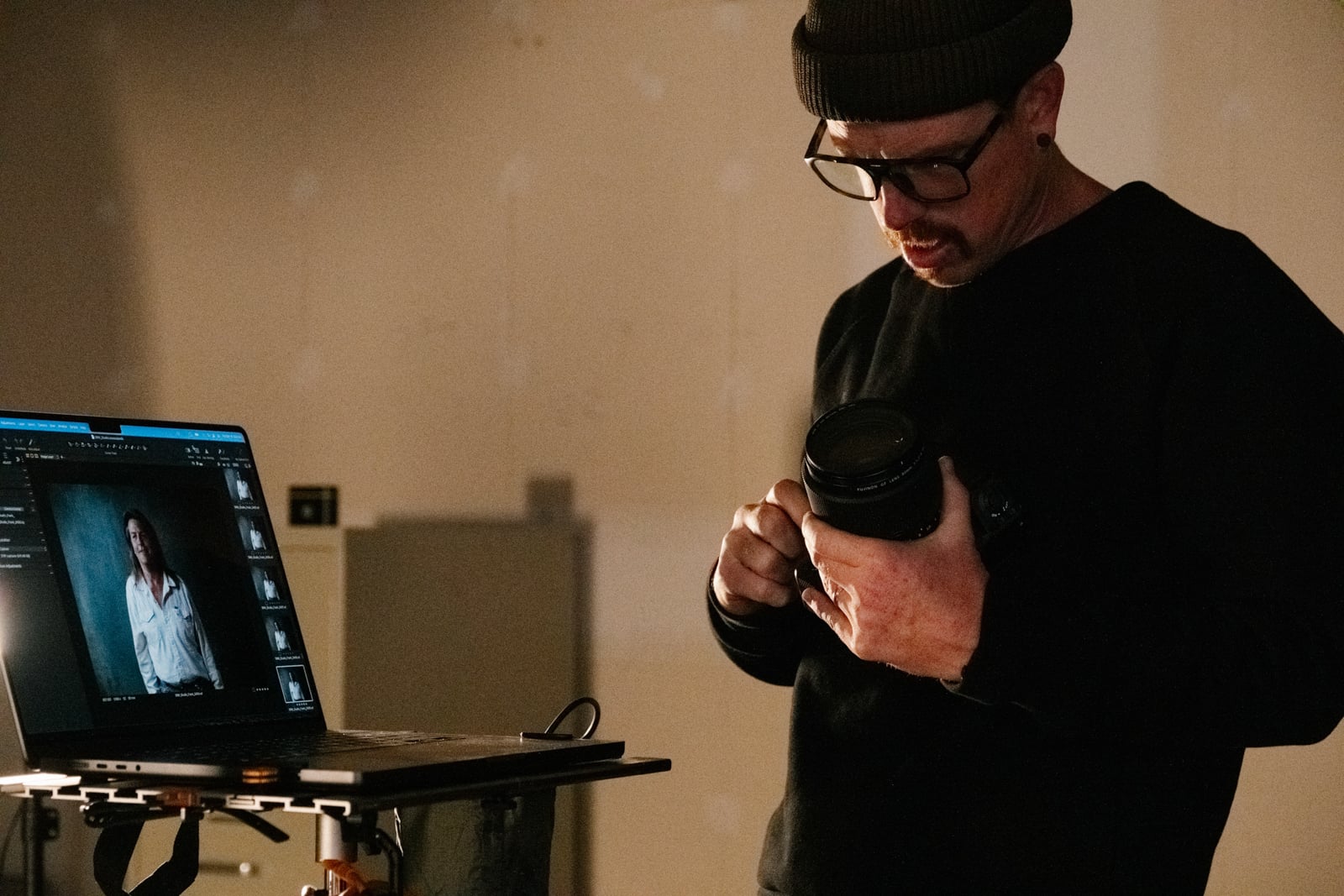 A man wearing glasses and a beanie examines a camera lens while standing next to a laptop displaying portrait photo editing software with a woman's image on the screen.